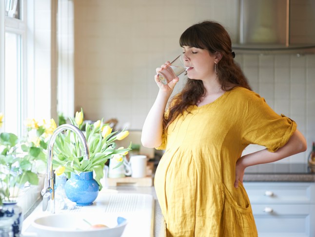 Pregnant woman drinking glass of water in kitchen at home.