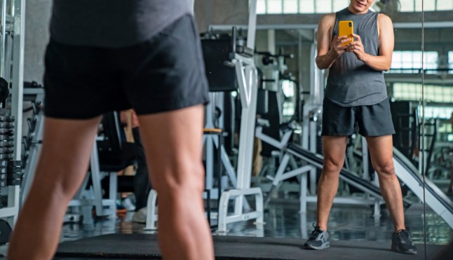 Strong Asian athletic man in sportswear using mobile phone taking selfie on mirror while workout exercise at fitness gym. Healthy male do sport training at sport club. Health care motivation concept.