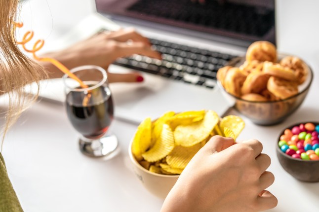 Girl works at a computer and eats fast food. Unhealthy food: chips, crackers, candy, waffles, cola. Junk food, concept.