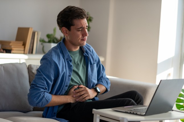 Young man sitting on the sofa at home working on his laptop, suffering from a stomach ache, holding his belly with his hands
